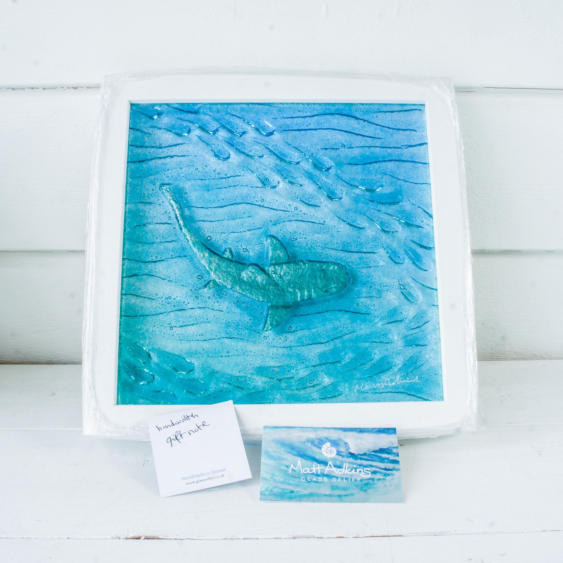 silver shimmering shark swimming in turquoise blue water with shoal of fish in textured fused glass in a white wooden frame 25x25cm (10")
