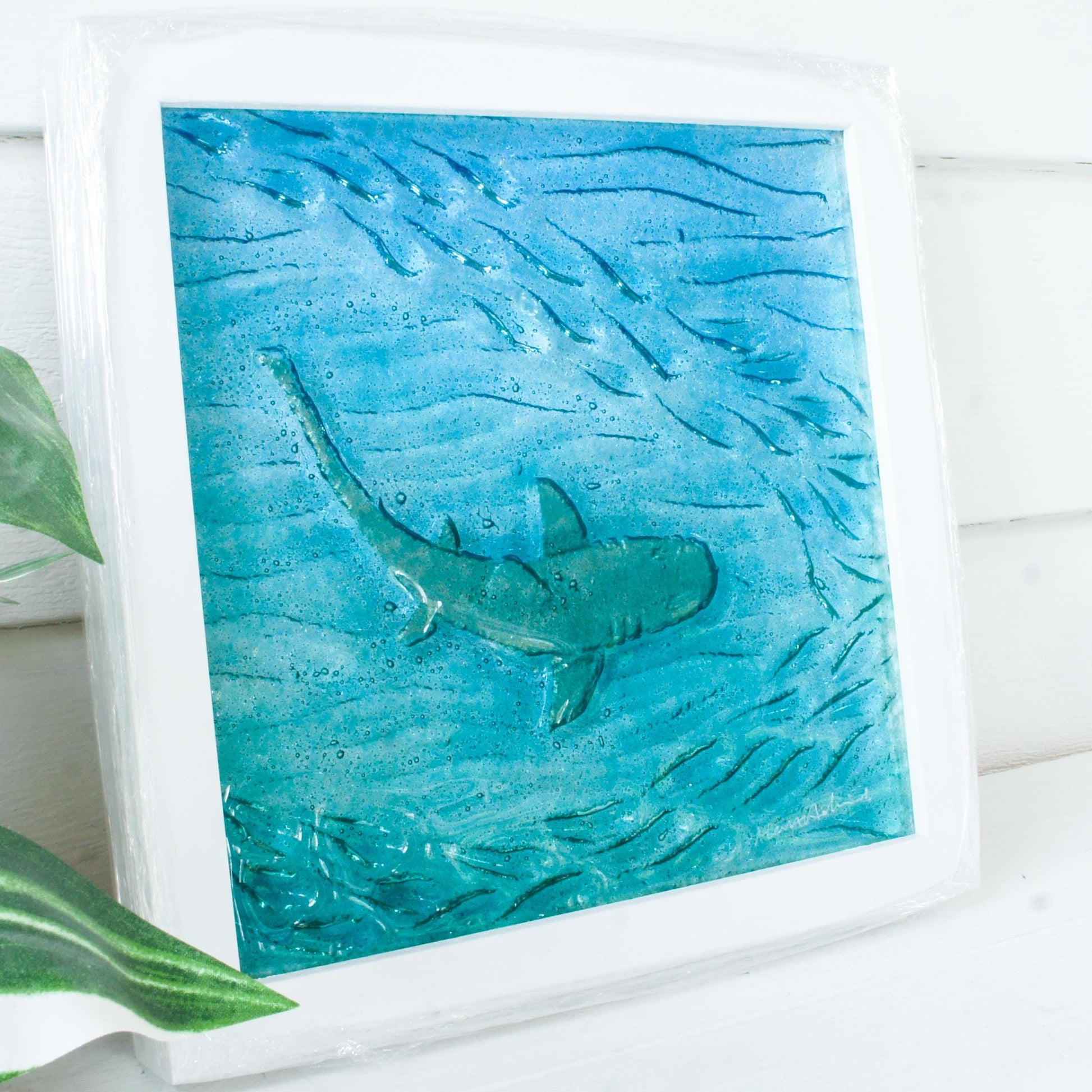 silver shimmering shark swimming in turquoise blue water with shoal of fish in textured fused glass in a white wooden frame 25x25cm (10")
