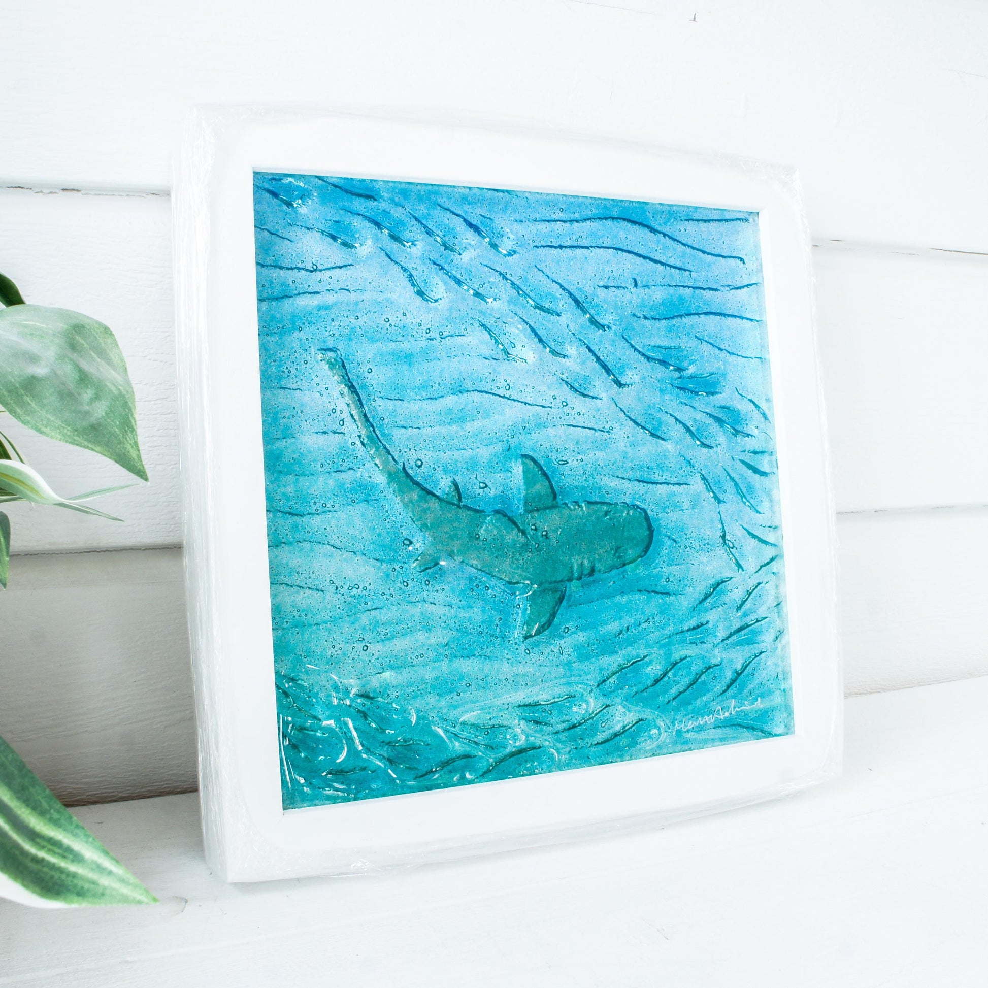 silver shimmering shark swimming in turquoise blue water with shoal of fish in textured fused glass in a white wooden frame 25x25cm (10")