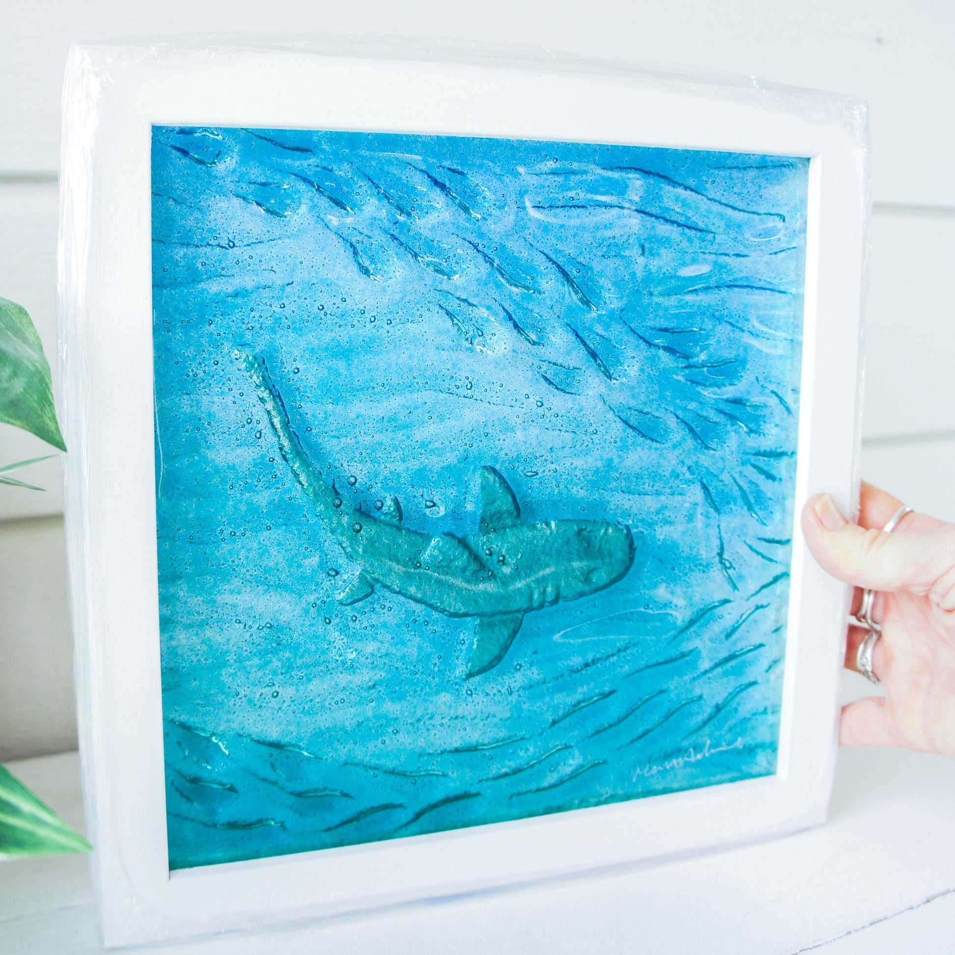 silver shimmering shark swimming in turquoise blue water with shoal of fish in textured fused glass in a white wooden frame 25x25cm (10")