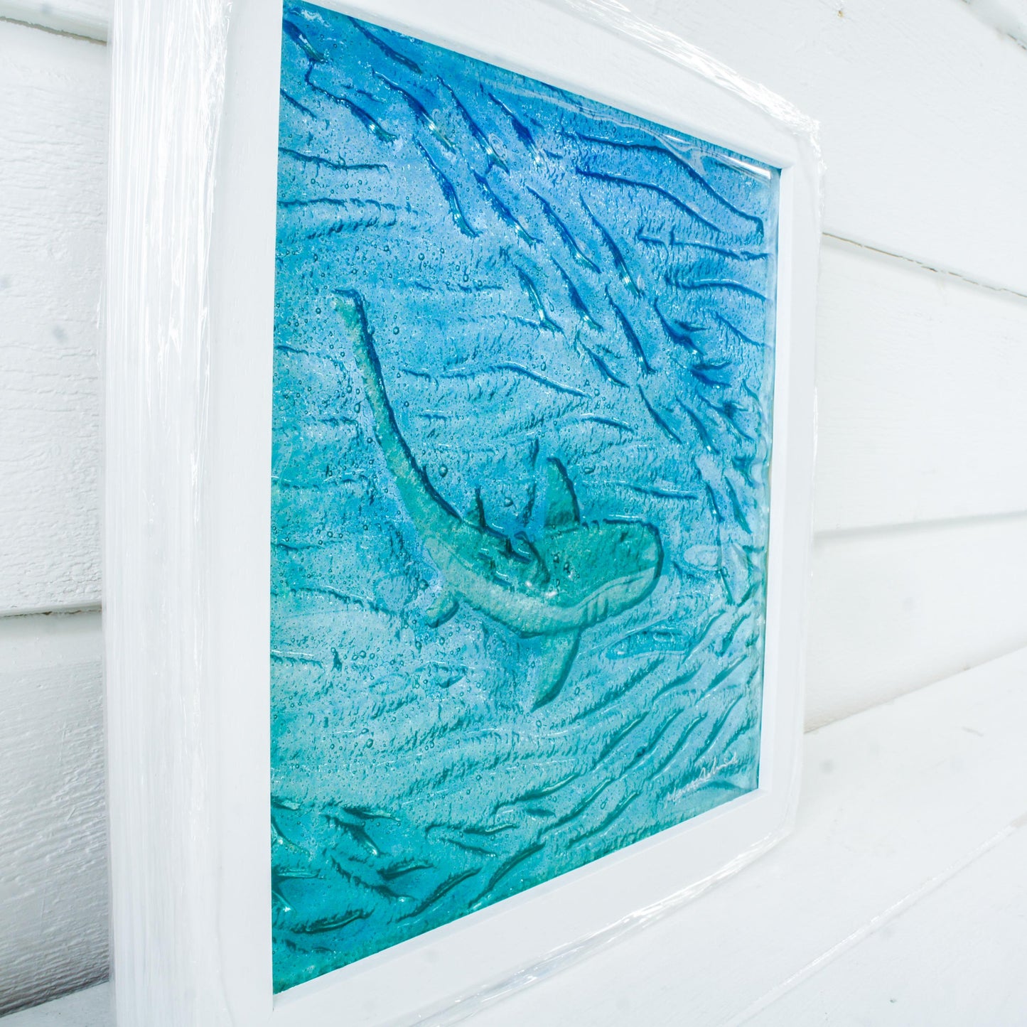 silver shimmering shark swimming in turquoise blue water with shoal of fish in textured fused glass in a white wooden frame 25x25cm (10")