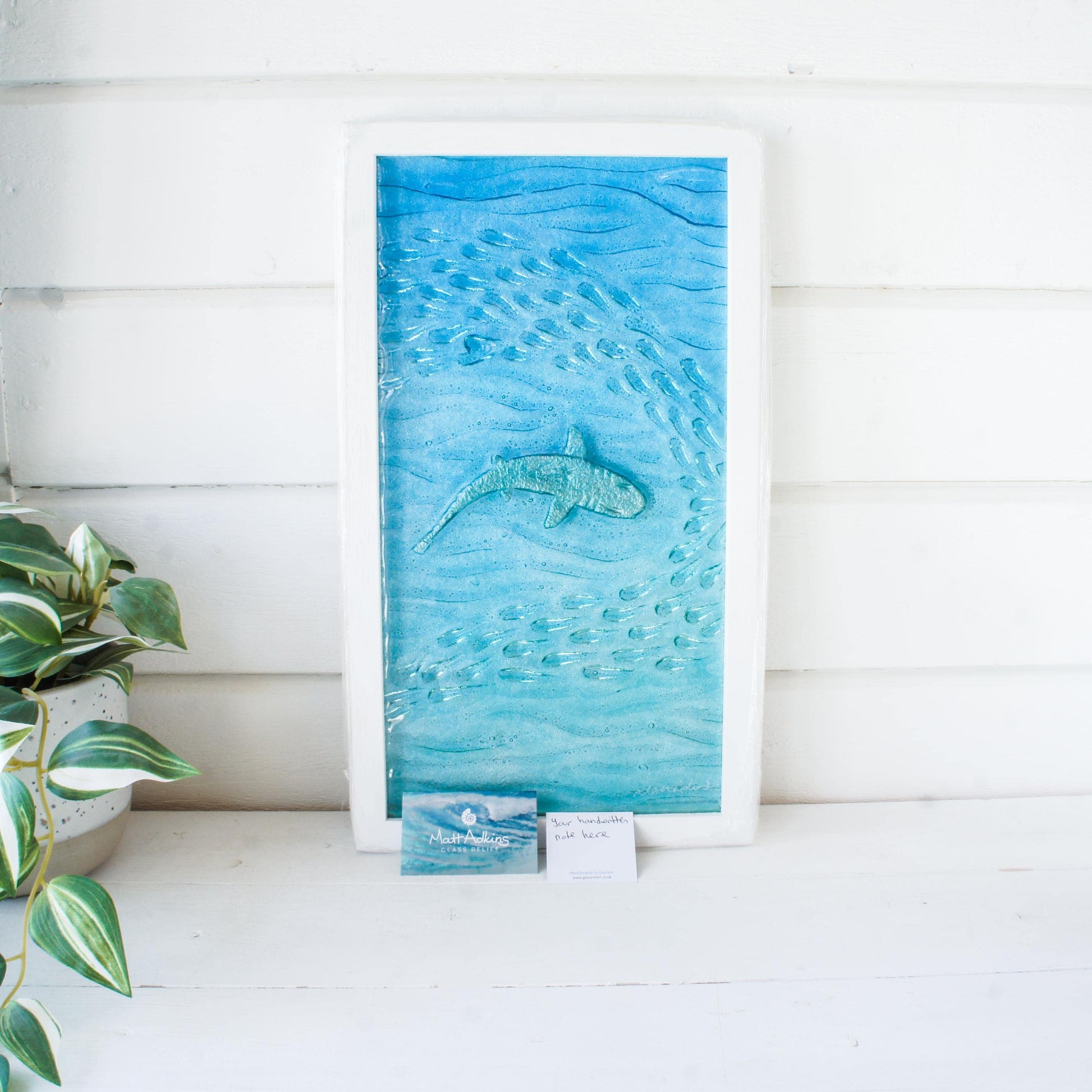 shark painted in silver swimming sideway with fish in blue sea fused glass wall art in a white wooden frame 25x45cm with gift note and business card