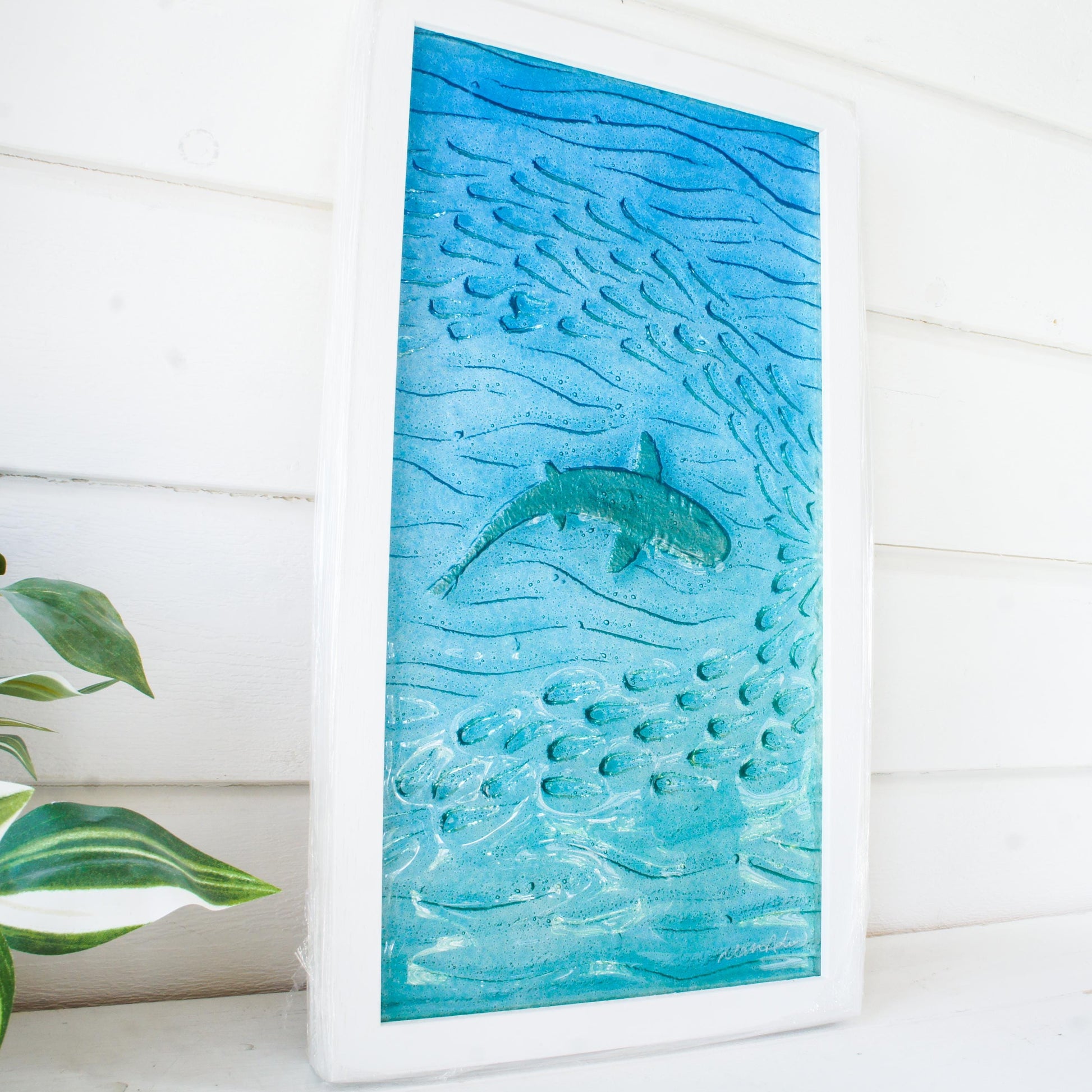 shark painted in silver swimming sideway with fish in teal sea fused glass wall art in a white wooden frame 25x45cm view from side